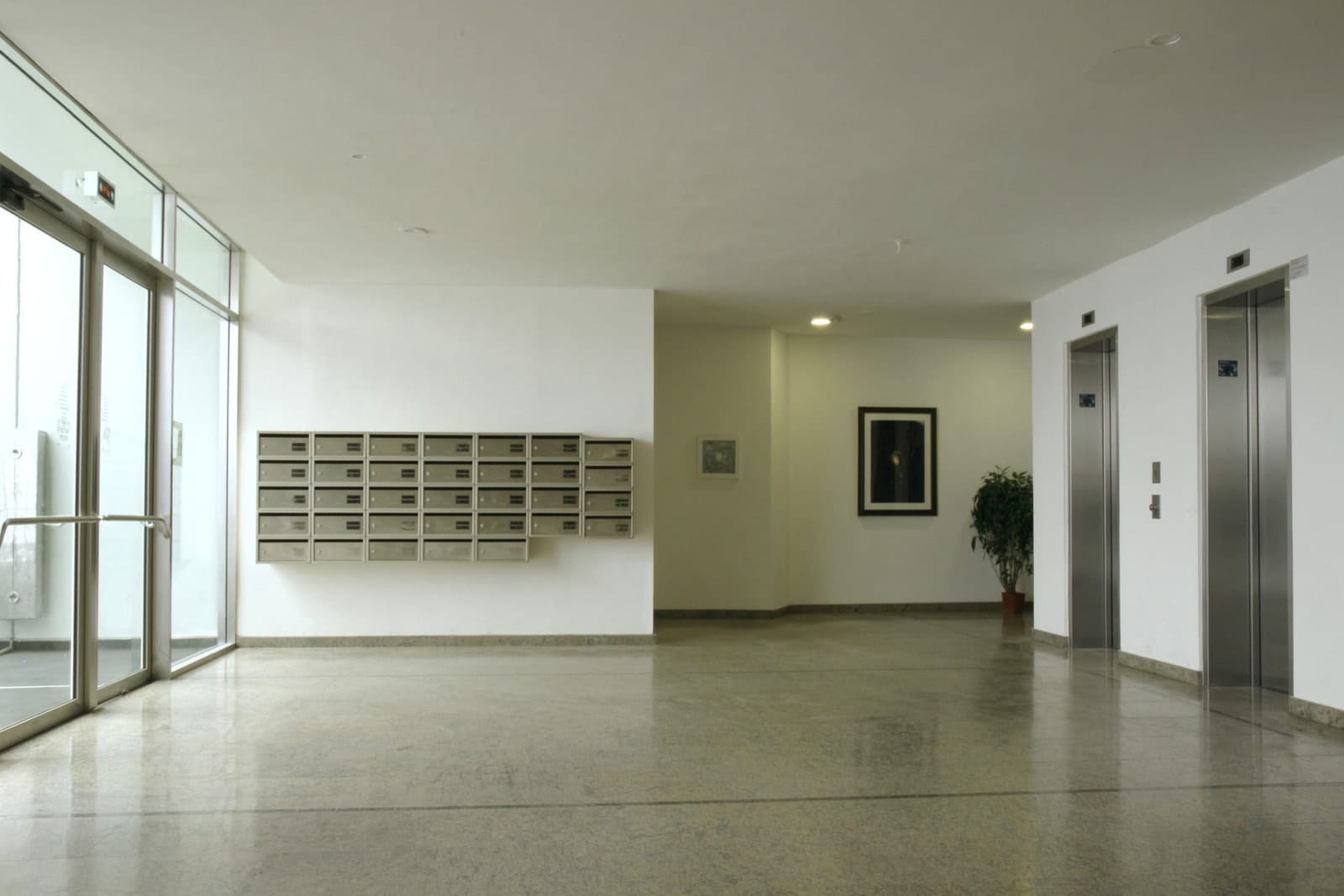 An empty apartment building lobby with a polished stone floor, glass doors and lift entrances.
